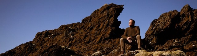 Alfred Gislason in braunem Pullover sitzt auf Felsen, umgeben von steiniger Landschaft unter klarem blauem Himmel.