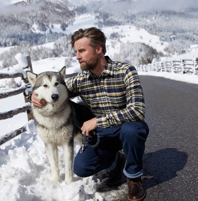 Mann hockt neben einem Husky in einer Schnee Landschaft.