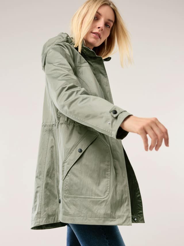 Eine Frau trägt eine hellgrüne, längere Jacke mit Kapuze und großen Taschen und bewegt einen Arm leicht nach vorn.