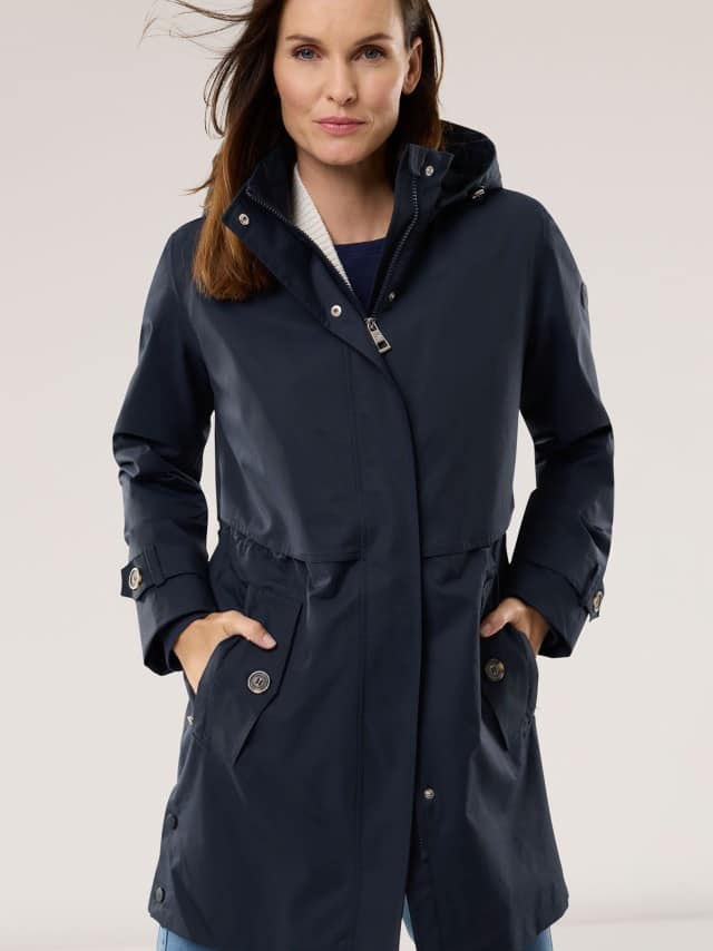 Eine Frau trägt eine dunkelblaue, lange Jacke mit Kapuze und hat die Hände in den Taschen.