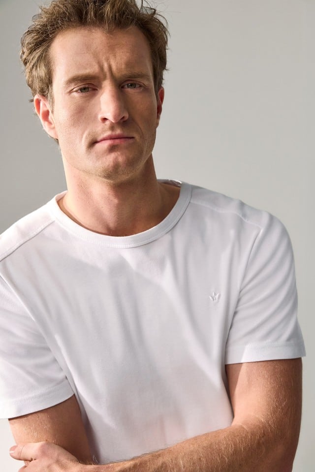 Ein Mann trägt ein schlichtes weißes T‑Shirt und blickt ernst in die Kamera.