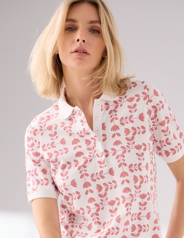 Eine Frau trägt ein Extraglatt-Poloshirt mit rosa Blumenmuster auf weißem Hintergrund.