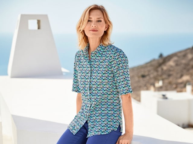 Frau mit einer gemusterten Hemdbluse sitzt auf einer weißen Mauer, im Hintergrund sieht man das Meer.