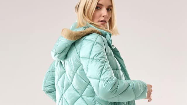 Eine Frau trägt eine mintgrüne, gesteppte Winterjacke mit Kapuze und beigefarbenem Futter. Der Stil wirkt modern und lässig.
