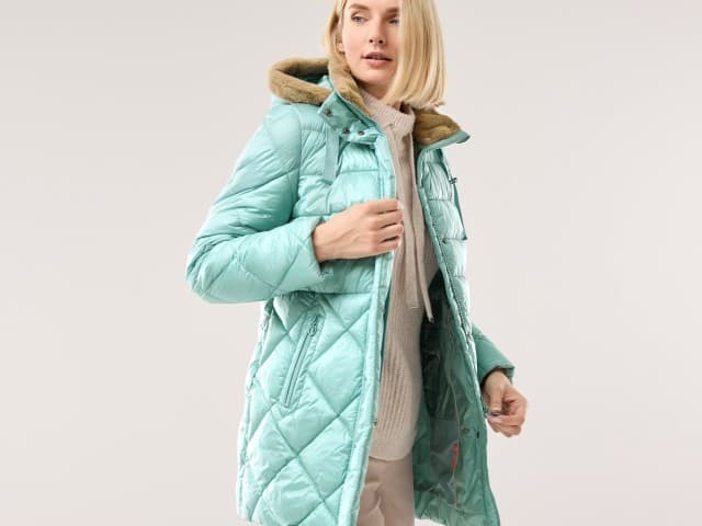 Eine Frau trägt eine lange, mintgrüne Steppjacke mit Kapuze und beigefarbenem Innenfutter. Darunter ist ein beiger Strickpullover zu sehen, der Look wirkt gemütlich und elegant.