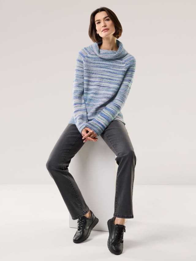 Eine Frau trägt einen gestreiften, lockeren Pullover in Blau- und Grüntönen mit weitem Kragen. Dazu kombiniert sie graue Jeans und schwarze Sneaker für einen gemütlich-modernen Look.