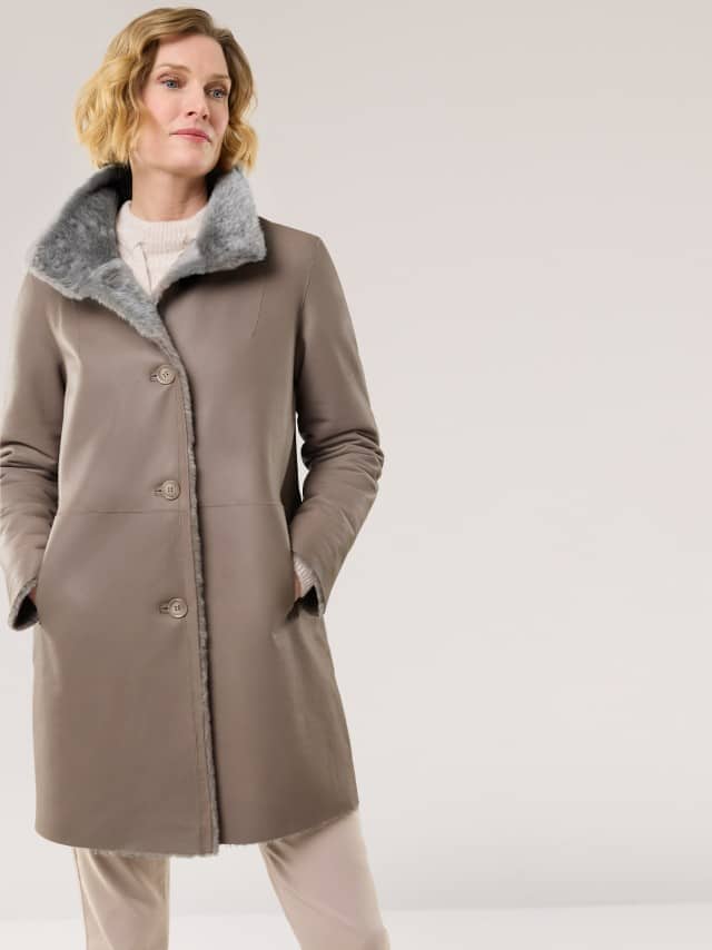 Eine Frau trägt einen knie­langen, taupefarbenen Mantel mit grauem Fellkragen. Der Look ist elegant und warm, ideal für die kalte Jahreszeit.