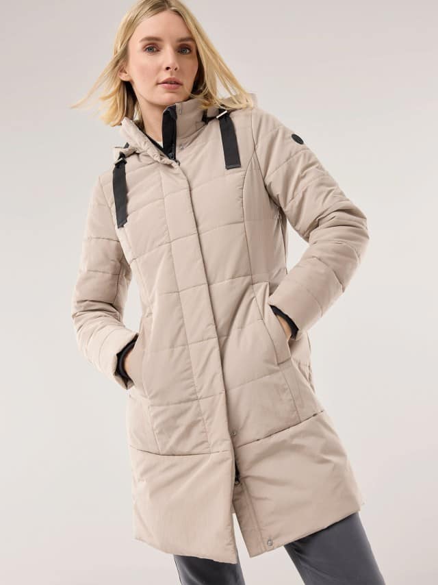 Eine Frau trägt eine hellbeige, gesteppte Winterjacke mit Kapuze und schwarzen Riemendetails. Der Stil wirkt sportlich und leger zugleich.