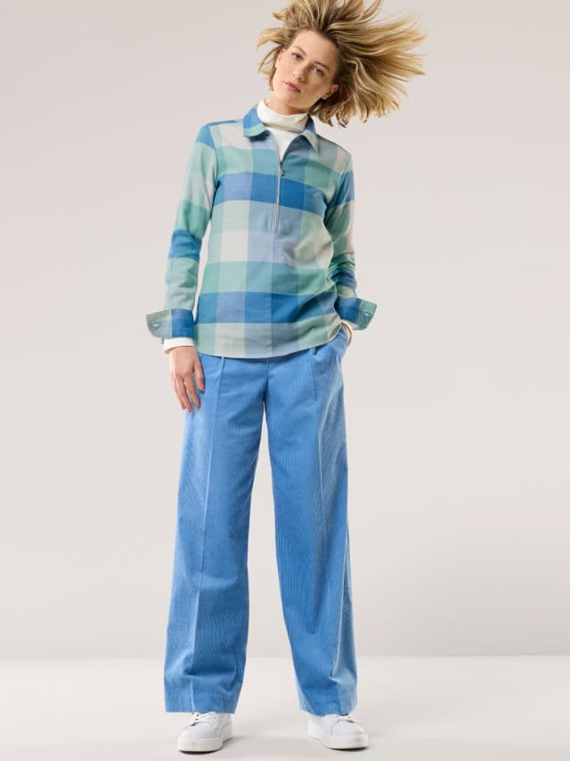 Eine Frau trägt ein kariertes Oberteil in Blau- und Grüntönen über einem weißen Rollkragen. Dazu kombiniert sie weite, hellblaue Cordhosen und weiße Sneaker für einen modernen, frischen Look.