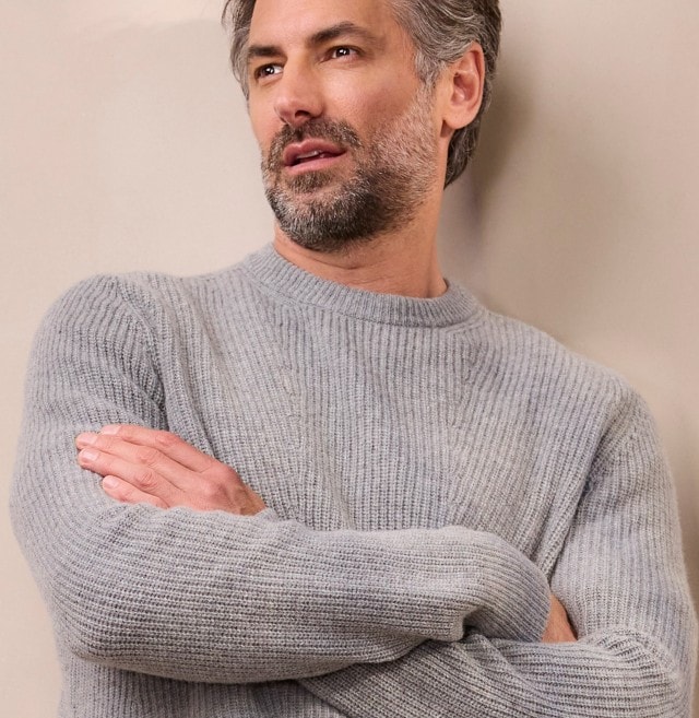 Ein Mann in einem grauen Strickpullover lehnt lässig an einer hellen Wand und schaut zur Seite.