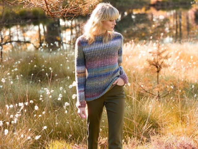 Frau in einem gestreiften Strickpullover in Herbsttönen und dunkler Hose steht in einem Feld bei Sonnenuntergang.