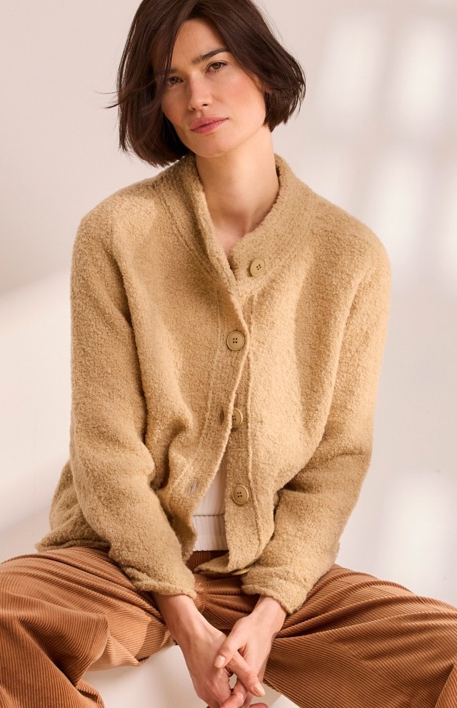 Frau in einer hellbraunen Strickjacke über einem weißen Pullover und einer braunen Cordhose sitzt in einem hellen Raum und schaut in die Kamera.