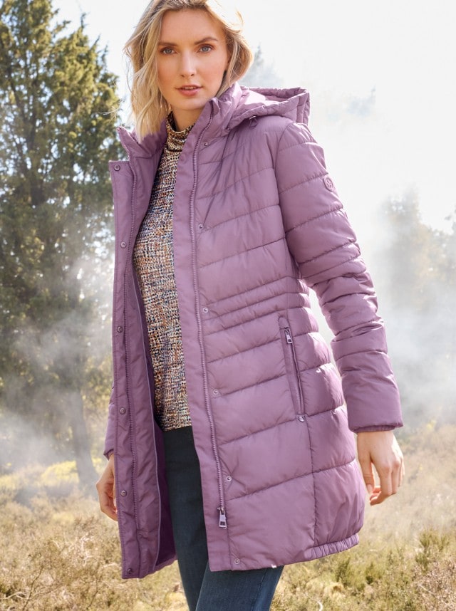 Frau in einem gemusterten Rollkragenpullover, Jeans und lila-farbenem Steppmantel steht in einem Feld und schaut in die Kamera.