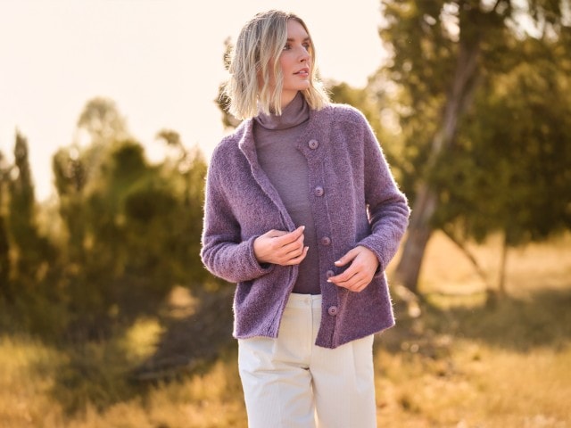 Frau in einem gemusterten Rollkragenpullover, lila-farbener Strickjacke und weißer Hose steht vor einem herbstlichen Hintergrund und schaut zur Seite.