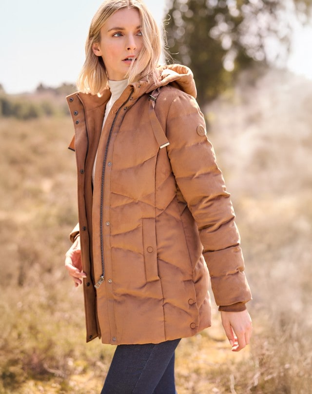 Frau in einem weißen Rollkragenpullover, einer hellbraunen Steppjacke und Jeans steht in einem Feld und schaut zur Seite.