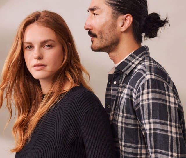 Eine Frau in einem schwarzen Strickpullover und ein Mann in einem schwarz-weiß-karierten Flanellhemd stehen aneinander gelehnt vor einem neutralen Hintergrund. 