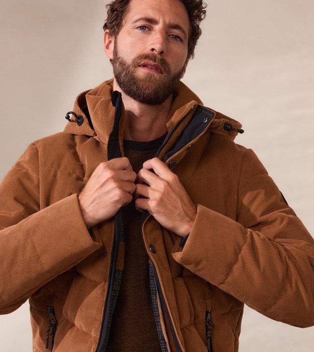 Eine Frau trägt eine dunkelblaue Jacke über einem blau-weiß gestreiften Pullover und eine rote Mütze. Ein Mann trägt eine braune Cord-Jacke mit Reißverschluss und Kapuze.