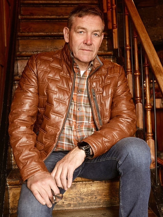 Alfred Gislason im Outfit Lammleder Leichtsteppjacke auf einer Treppe sitzend.