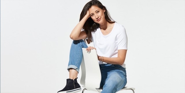 Frau in weißem T-Shirt und blauer Jeans sitzt auf einem Stuhl, lehnt den Kopf auf die Hand.