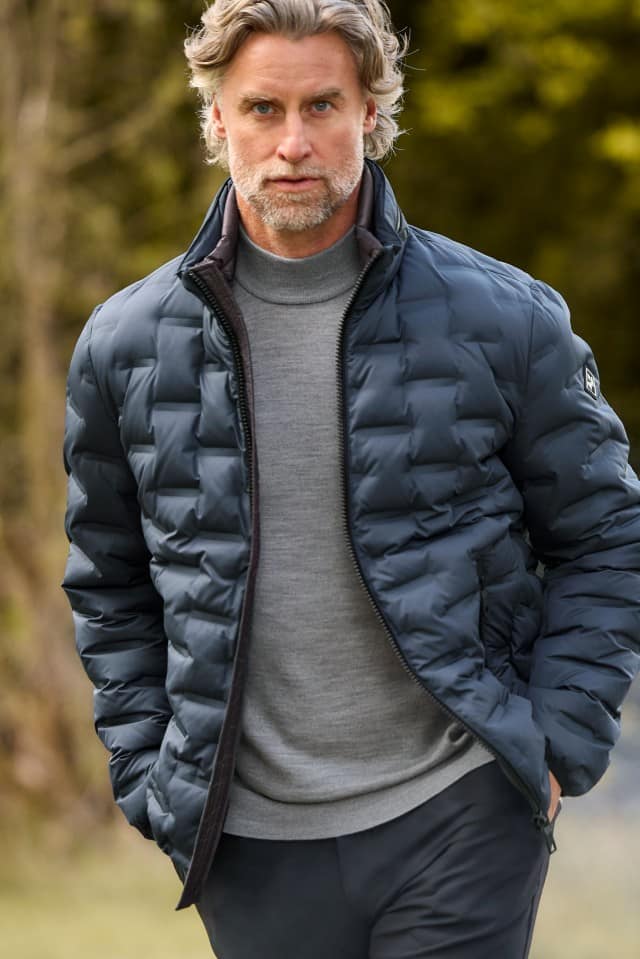 Ein Mann trägt eine dunkelblaue Steppjacke über einem grauen Pullover mit Rollkragen. Das Outfit wirkt sportlich-elegant und eignet sich gut für kühle Tage im Freien.