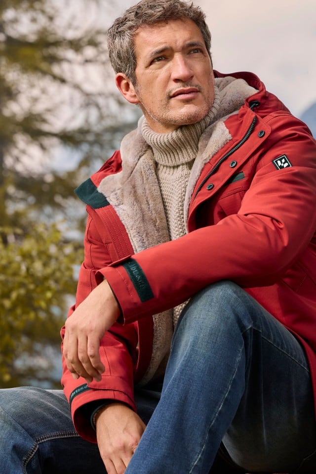 Ein Mann trägt eine rote Winterjacke mit Fellfutter über einem dicken, beigen Rollkragenpullover. Das Outfit wirkt robust, warm und perfekt für Outdoor-Aktivitäten.