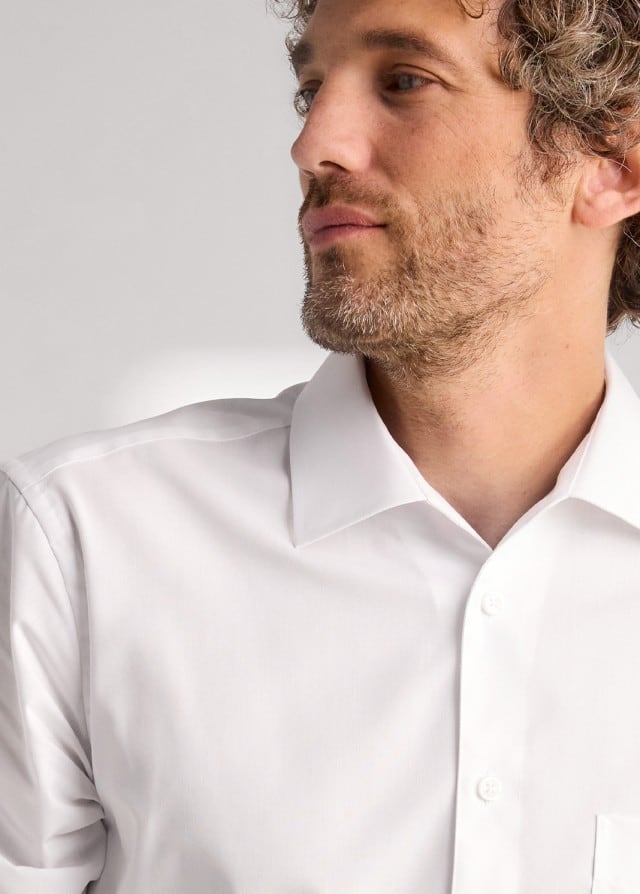 Ein Mann mit lockigem Haar, trägt ein weißes Hemd und schaut zur Seite.