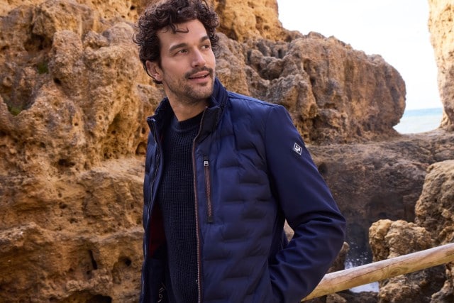Mann in einer dunkelblauen Steppjacke über einem dunklen Strickpullover steht vor Felsen und schaut lächelnd zur rechten Seite.