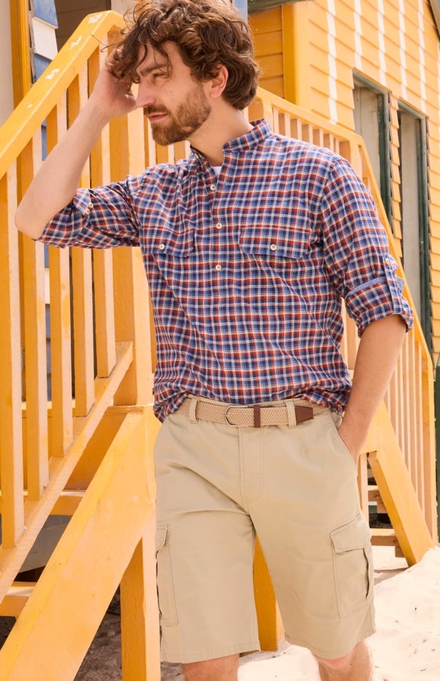 Ein Mann trägt ein blau-rot kariertes Langarmhemd und eine beige Cargoshorts mit Gürtel und steht vor einem gelben Holzhaus an einer Treppe.