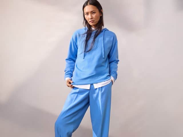 Eine Frau trägt einen hellblauen Hoodie über einer weißen Bluse und eine dazu passende hellblaue Hose.