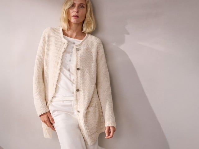 Eine Frau trägt eine cremefarbene Strickjacke über einem weißen Outfit und lehnt an einer Wand.