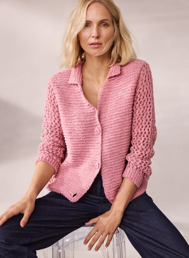 Eine Frau trägt eine rosafarbene Strickjacke mit durchbrochenen Ärmeln und sitzt auf einem transparenten Hocker.