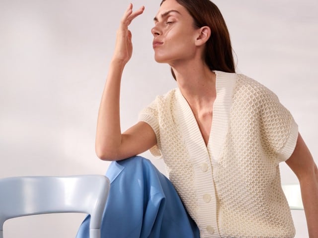 Eine Person sitzt auf einem Stuhl und trägt eine cremefarbene Strickjacke sowie eine hellblaue Hose, während sie mit geschlossenen Augen eine Hand vor das Gesicht hält.