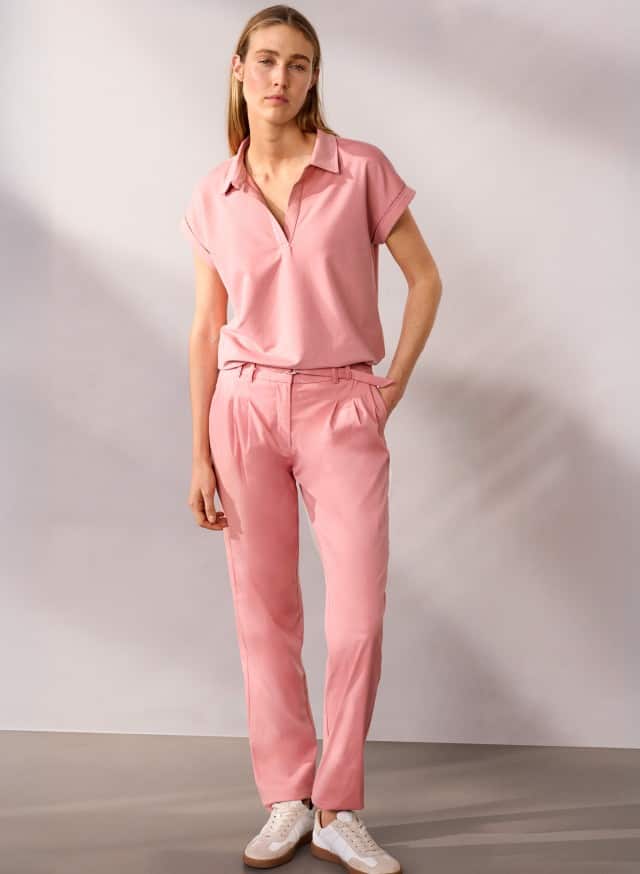 Eine Person steht vor einer neutralen Wand und trägt ein rosafarbenes Oberteil, eine passende rosafarbene Hose und weiße Sneaker.