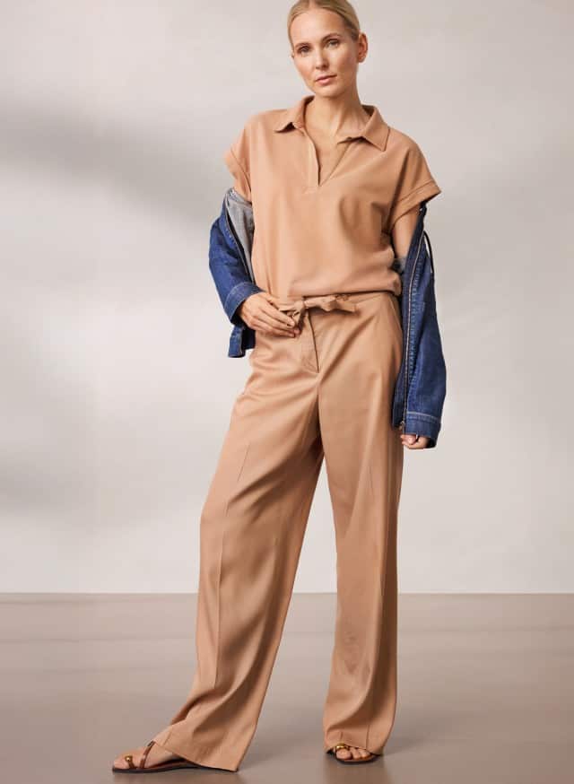 Eine Person steht vor einer neutralen Wand und trägt ein beigefarbenes Outfit mit weiter Hose und Kurzarmtop, kombiniert mit einer halb übergezogenen Jeansjacke und offenen Sandalen.