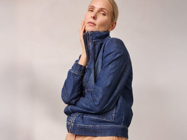 Eine Person trägt eine hochgeschlossene dunkelblaue Jeansjacke und berührt mit einer Hand ihr Gesicht.