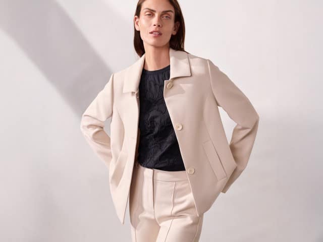 Eine Person steht vor einer neutralen Wand und trägt einen hellen Anzug mit Jacke und Hose, kombiniert mit einem dunklen Oberteil.