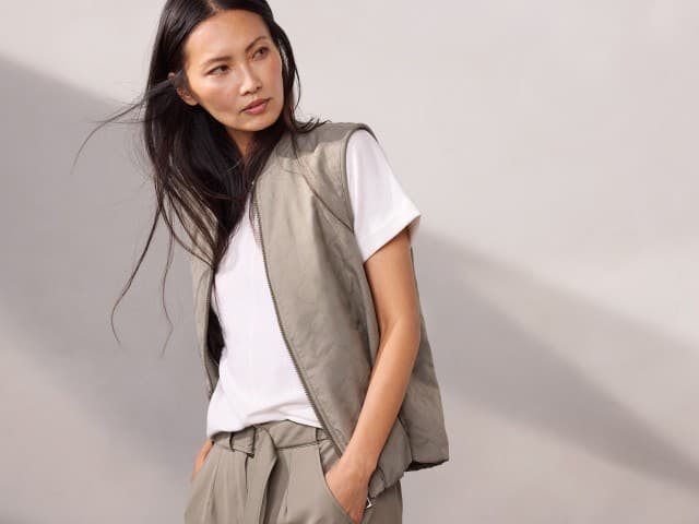 Eine Person trägt ein weißes T‑Shirt, eine leichte gesteppte Weste und eine locker sitzende beigefarbene Hose, während sie zur Seite blickt.