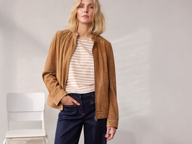 Eine Person steht vor einer neutralen Wand und trägt eine braune Jacke, ein gestreiftes Oberteil und eine dunkelblaue Jeans.