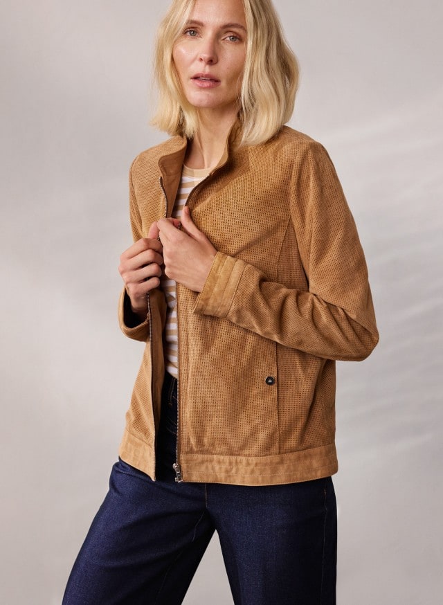 Eine Person trägt eine hellbraune Jacke über einem gestreiften Oberteil und eine dunkelblaue Jeans, während sie die Jacke mit beiden Händen hält.