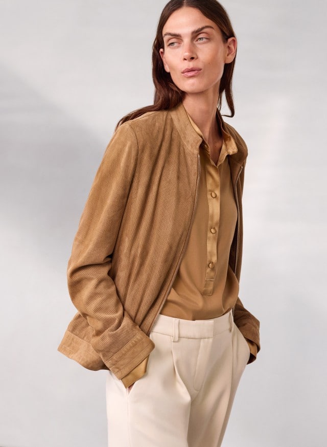 Eine Person trägt eine hellbraune Bluse, eine passende braune Jacke und eine helle Stoffhose und blickt zur Seite.