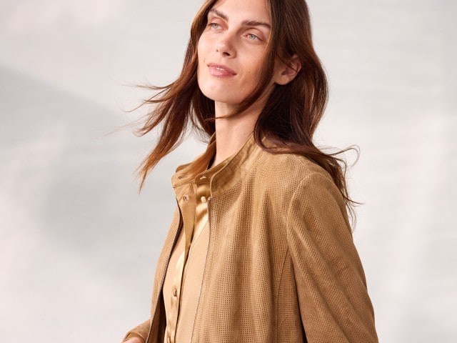 Eine Person trägt eine hellbraune Bluse und eine passende perforierte Jacke und blickt leicht lächelnd zur Seite.