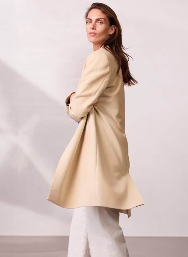 Eine Person trägt einen langen, hellen Mantel über einer weiten, hellen Hose und blickt über die Schulter zur Seite.