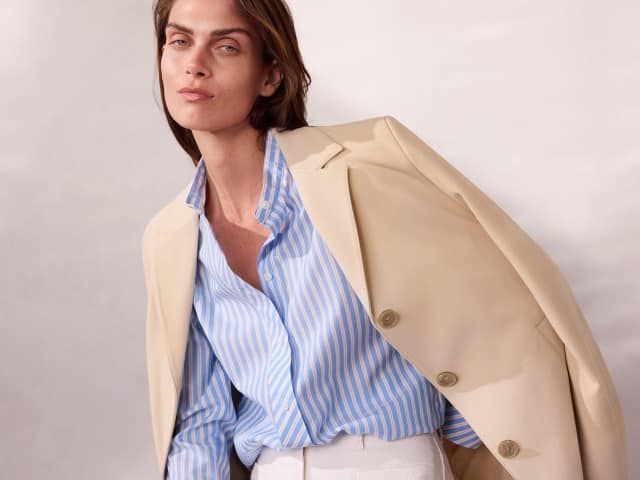 Eine Person trägt eine hellbeige Jacke über einer hellblau‑weiß gestreiften Bluse und einer weißen Hose und lehnt sich leicht nach vorn.