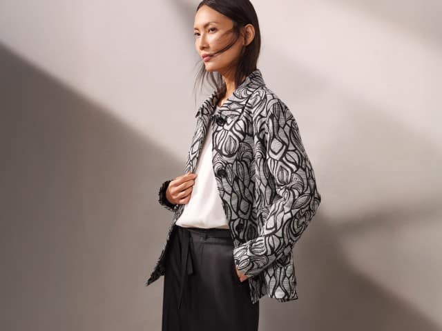 Eine Person trägt eine weiß‑schwarze gemusterte Jacke über einem weißen Oberteil und einer dunklen Hose und blickt zur Seite.