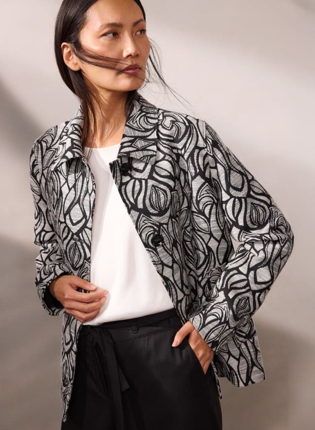 Eine Person trägt eine schwarz‑weiß gemusterte Jacke über einem weißen Oberteil und einer schwarzen Hose und blickt leicht zur Seite.