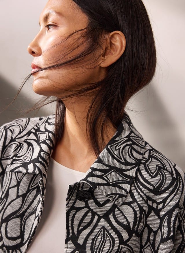 Eine Person trägt eine schwarz‑weiß gemusterte Jacke über einem weißen Oberteil und ist im Profil zu sehen.