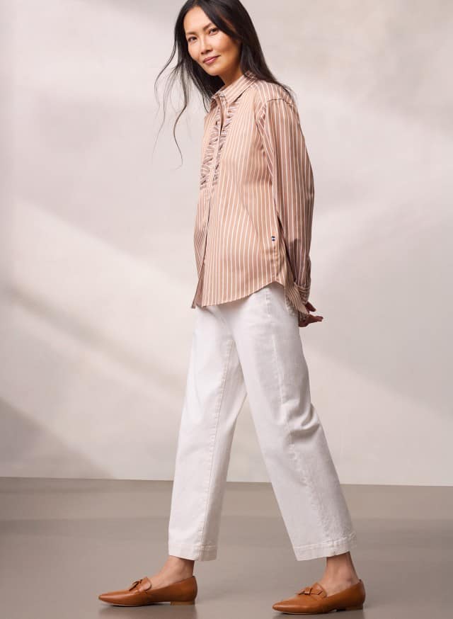 Eine Person trägt eine beigefarbene, gestreifte Bluse zu weißen, weiten Hosen und braunen Loafern und geht leicht zur Seite.