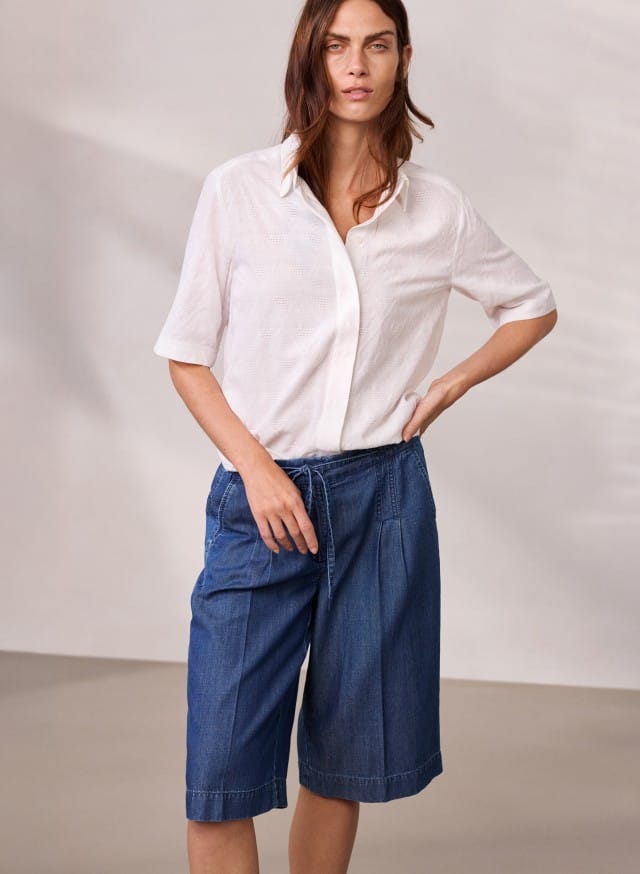 Eine Person trägt ein weißes Kurzarmhemd und knie­lange blaue Jeansshorts und steht vor einer neutralen Wand.