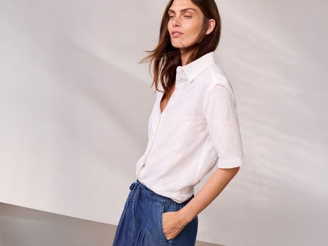 Eine Person trägt ein weißes Kurzarmhemd und blaue, knielange Hosen und blickt seitlich nach vorn.