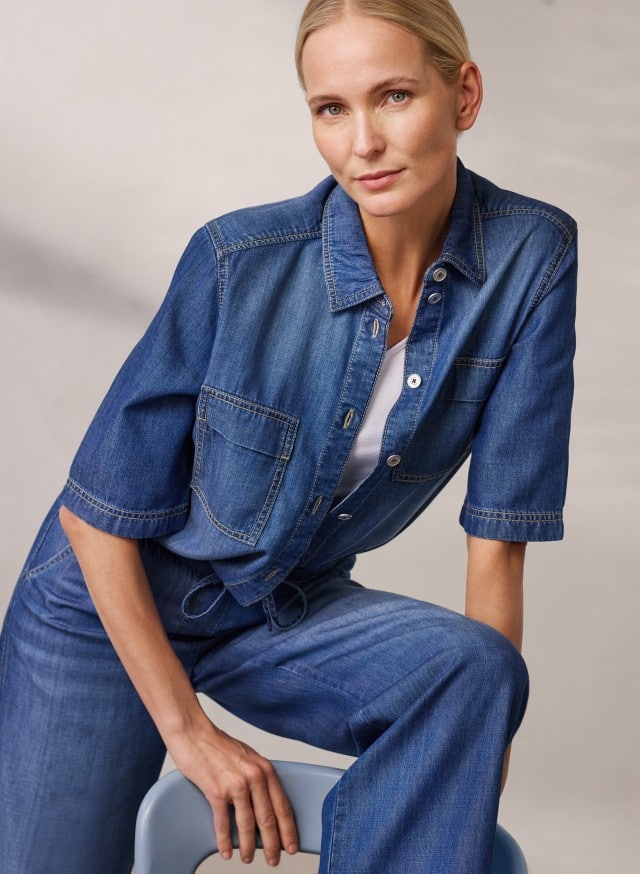 Eine Person trägt ein kurzärmliges Jeanshemd über einem weißen Top sowie eine passende weite Jeanshose und sitzt auf einem Stuhl.